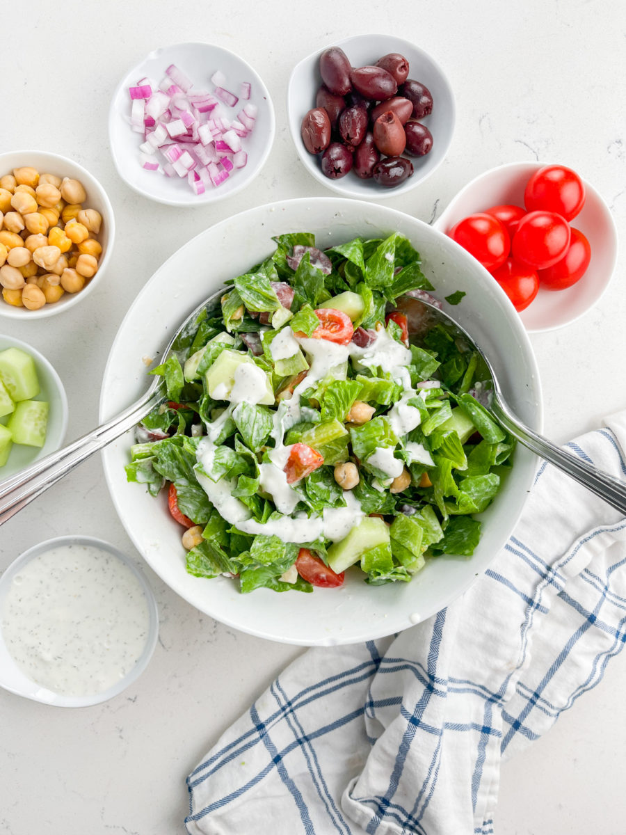 Greek Chopped Salad in a white bowl with dressing and serving spoons.